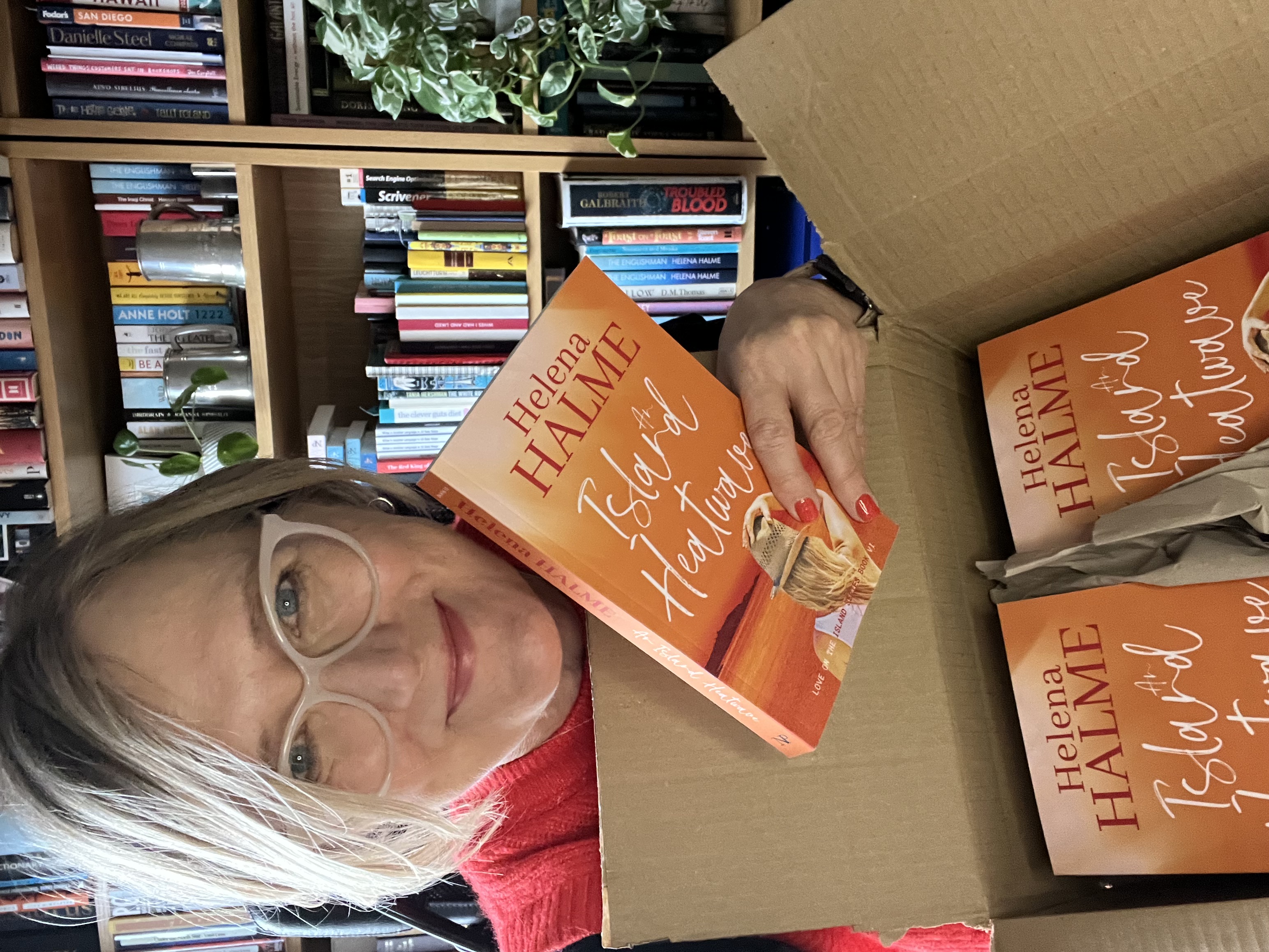 An image of Helena Halme smiling as she holds a box of paperback copies of AN ISLAND HEATWAVE.